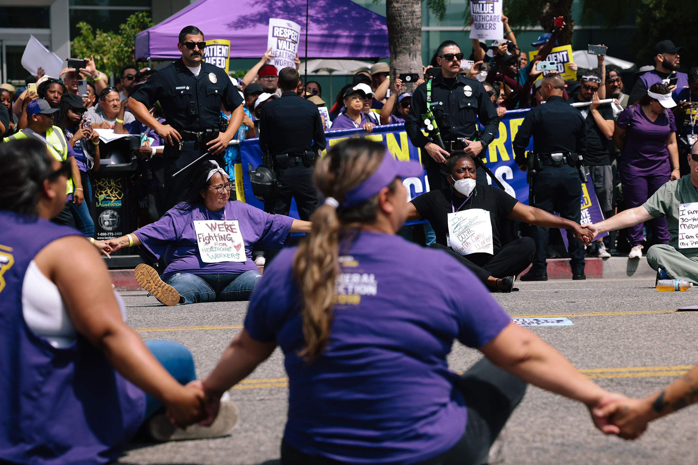 Healthcare workers, all dressed in purple shirts, sit in a street and hold hands in a circle as police officers stand watch in front of more purple-clad protesters holding a banner on a nearby sidewalk