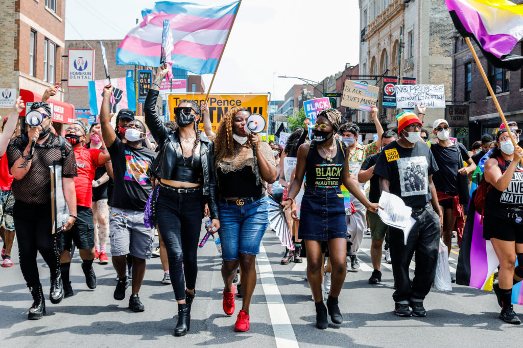 People march in the street, a central figure holding a megaphone to their mouth to chant, while holding Trans flags and signs expressing support for queer people