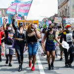 People march in the street, a central figure holding a megaphone to their mouth to chant, while holding Trans flags and signs expressing support for queer people