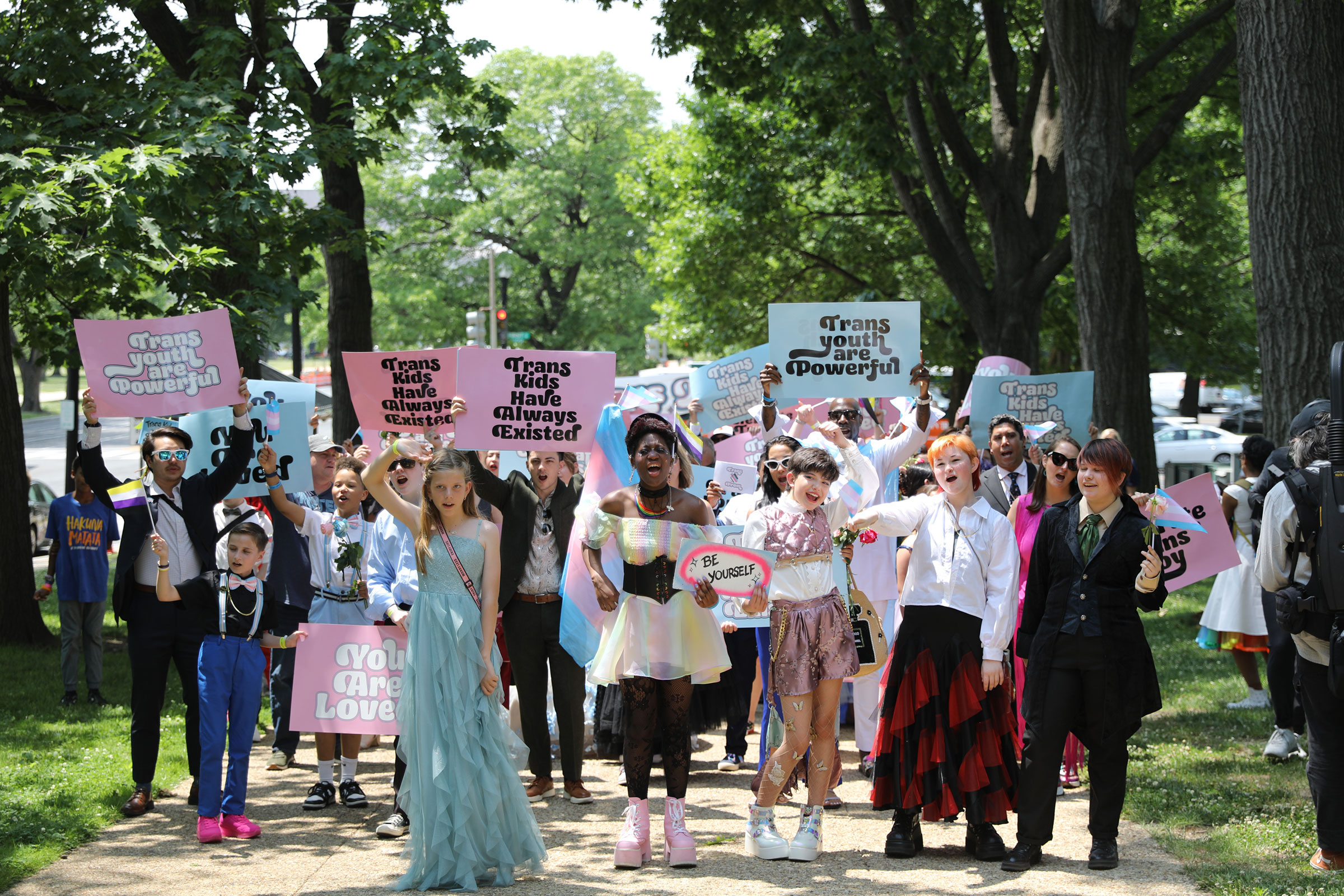 Trans youth, wearing colorful clothing and joyful expressions, march to the capitol holding signs reading