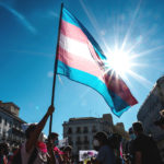 A person waves a trans flag in the sky during an outdoor protest