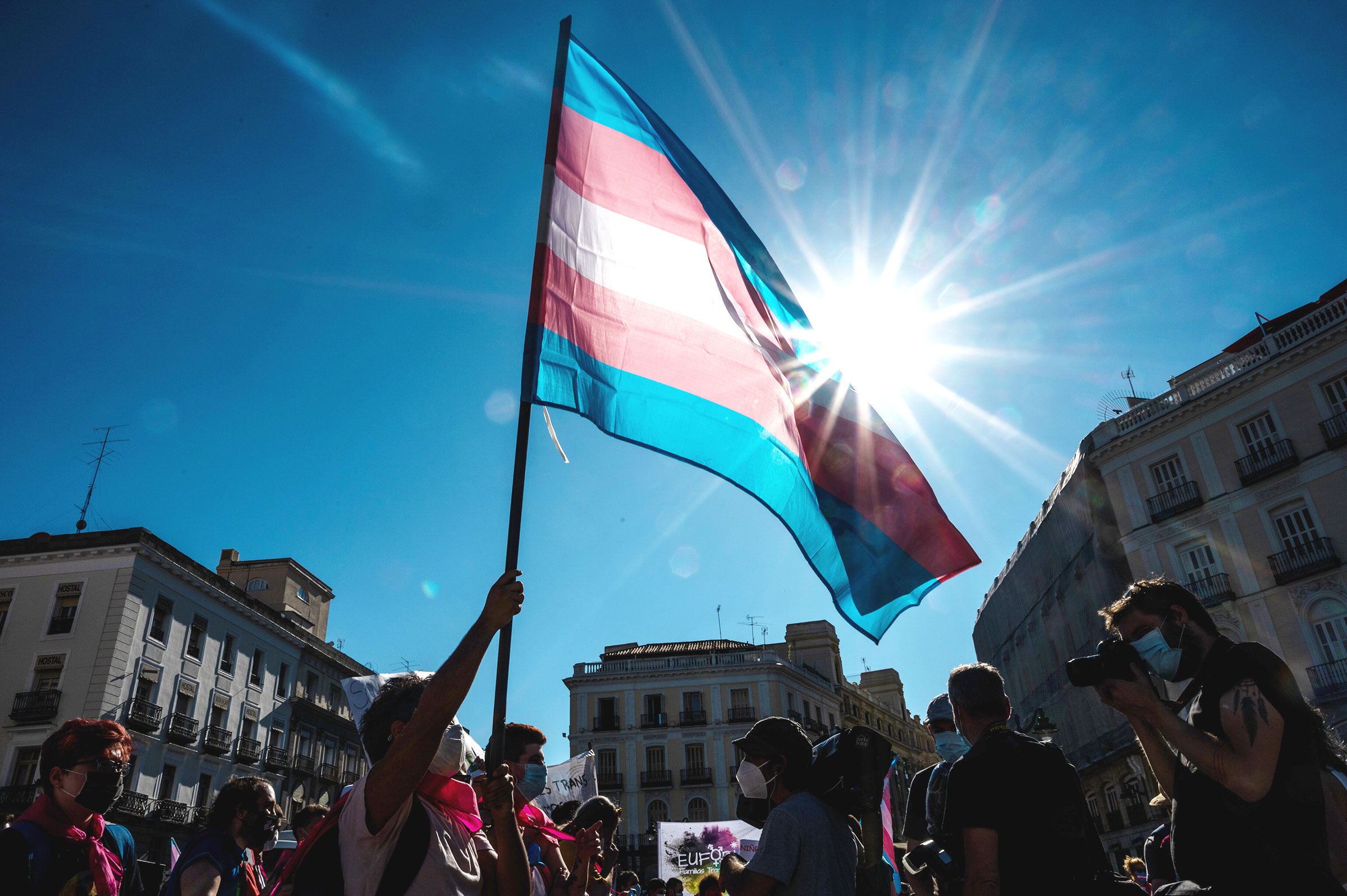 A person waves a trans flag in the sky during an outdoor protest