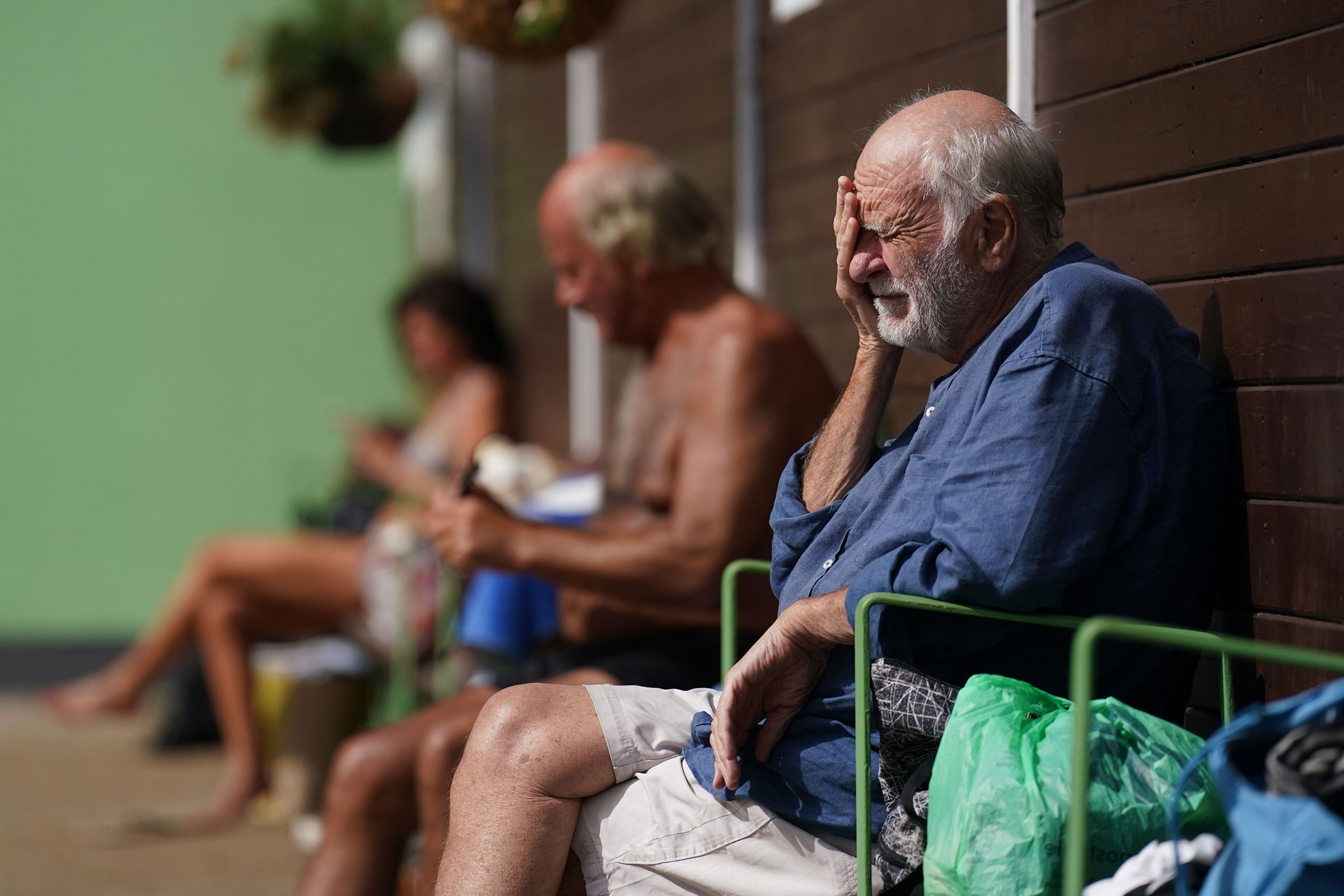 An elderly man sits against a wall and rests his face in his hands