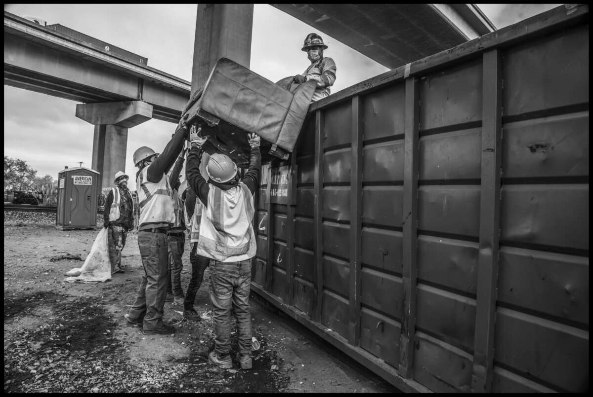 Des journaliers hissent un canapé abandonné dans une benne à ordures pour le mettre à la poubelle.  Sur le passage supérieur de l'autoroute, les camions quittent l'immense port d'Oakland.