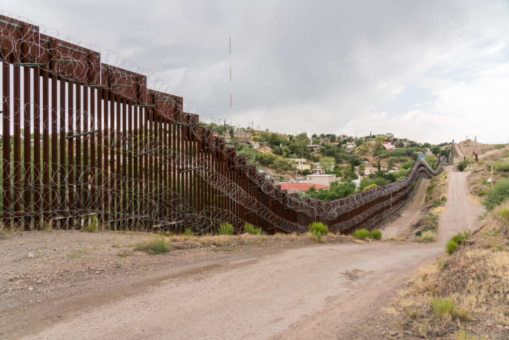 High steel wall with concertina wire, and the dirt road along where the border patrol truck drive on.