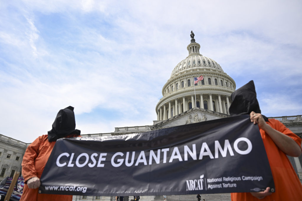 Two demonstrators in orange jumpsuits wearing black hoods carry a sign saying