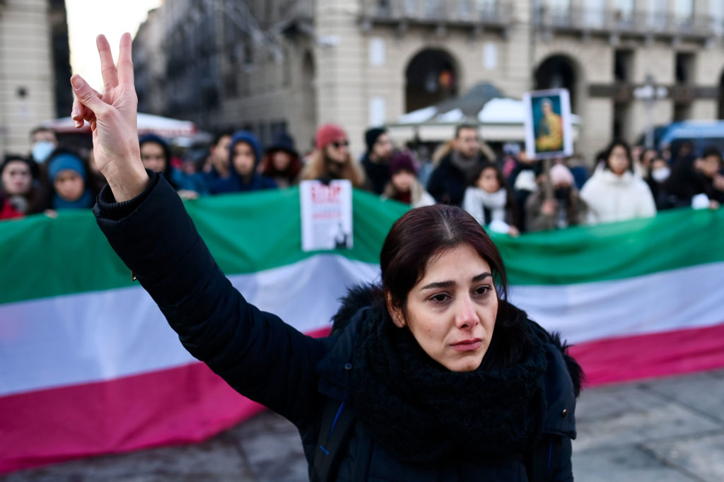A protester makes a peace sign while standing in front of others, who are holding a a banner painted to resemble the Iranian flag