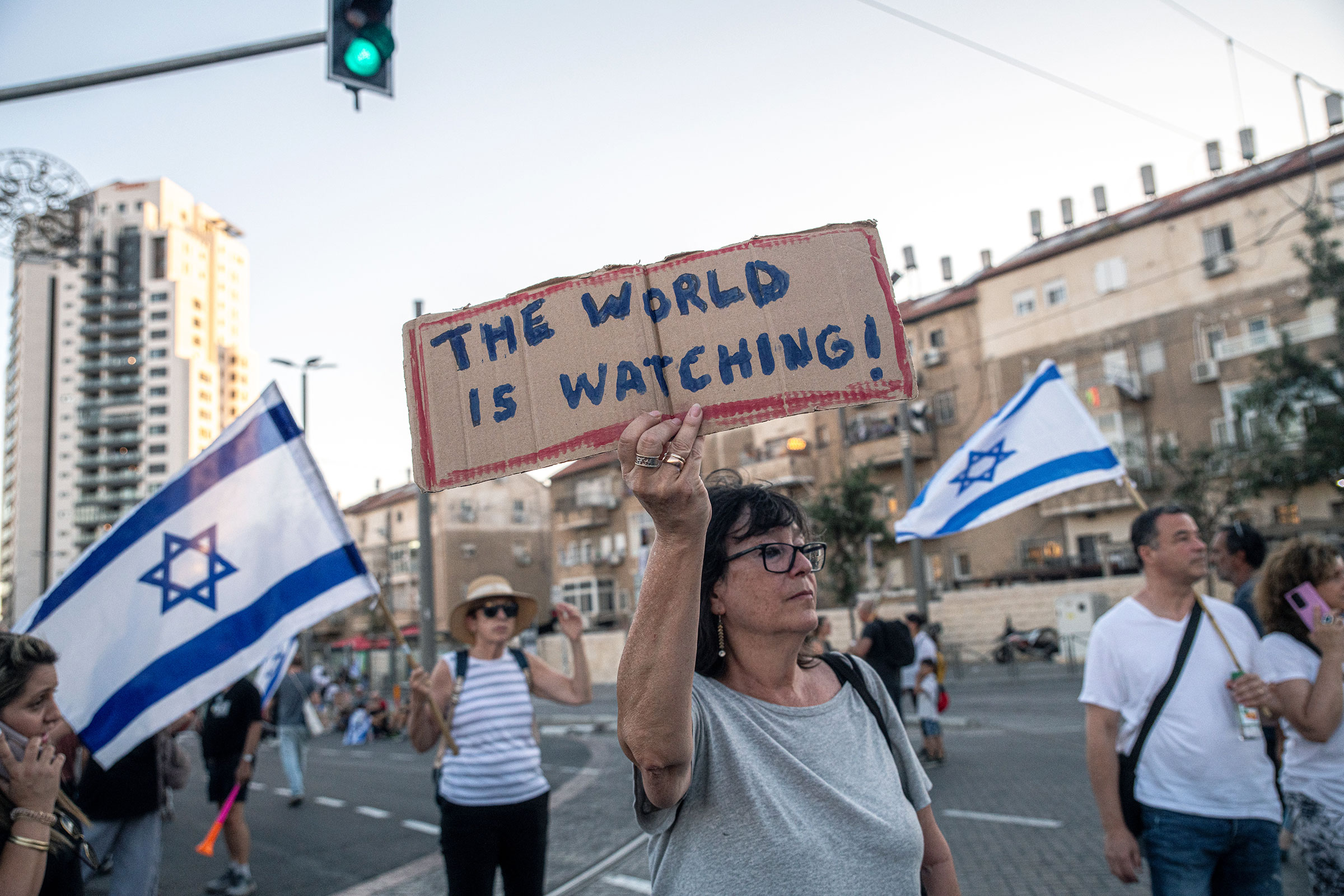 A woman, flanked by protesters behind her carrying the Israeli flag, holds a sign reading
