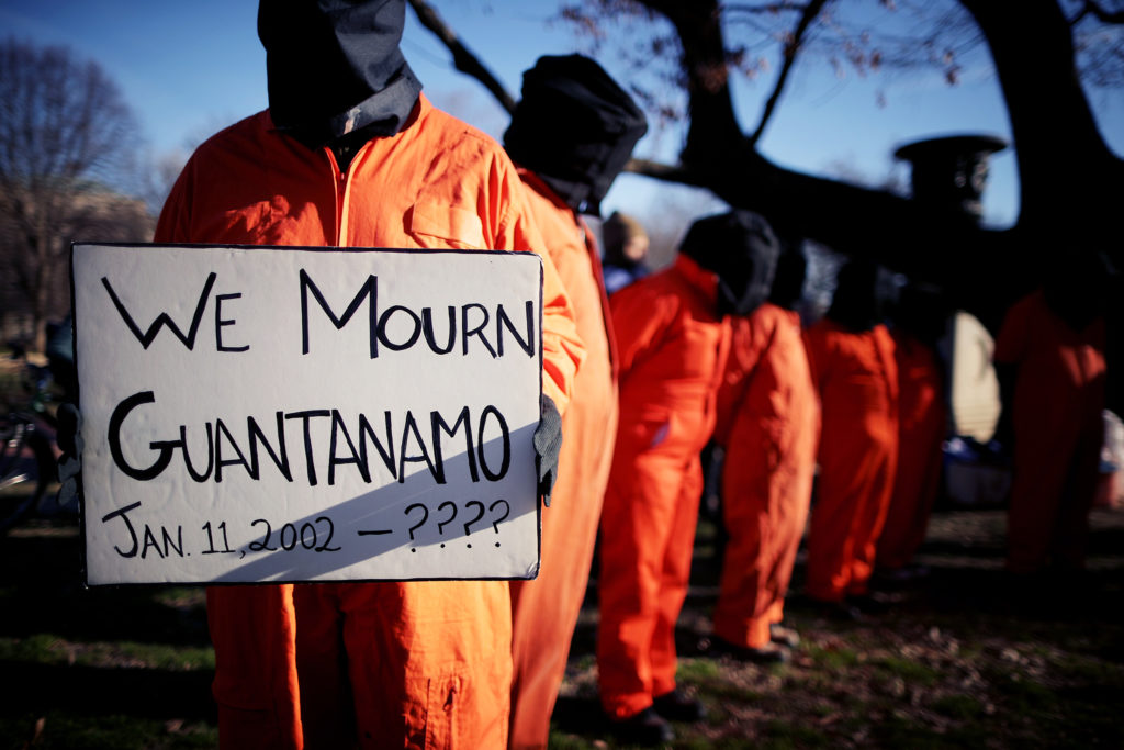 People in orange jumpsuits and hoods protest as one holds a sign reading