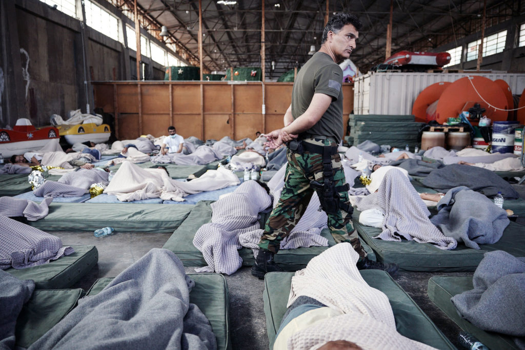 A man in military uniform patrolls refugees as they sleep on the floor of a warehouse