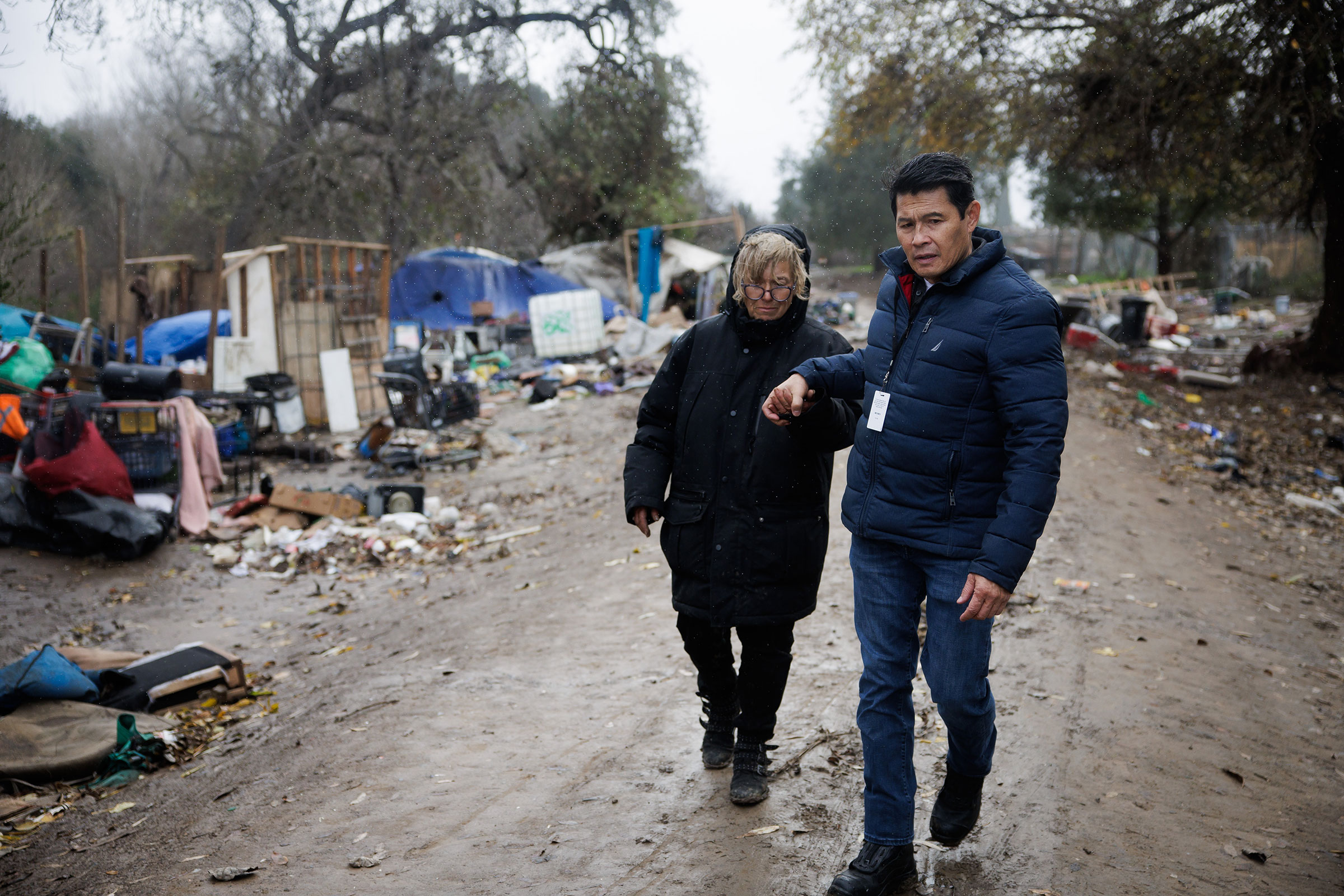 A man leads a woman by the hand through a homeless encampment in rainy weather