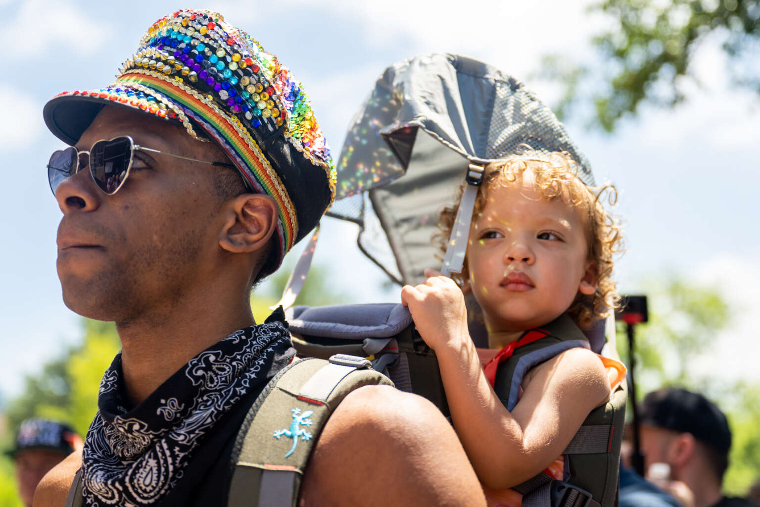 A parent wearing a rainbow hat carries their child on their back at a protest for LGBTQ rights