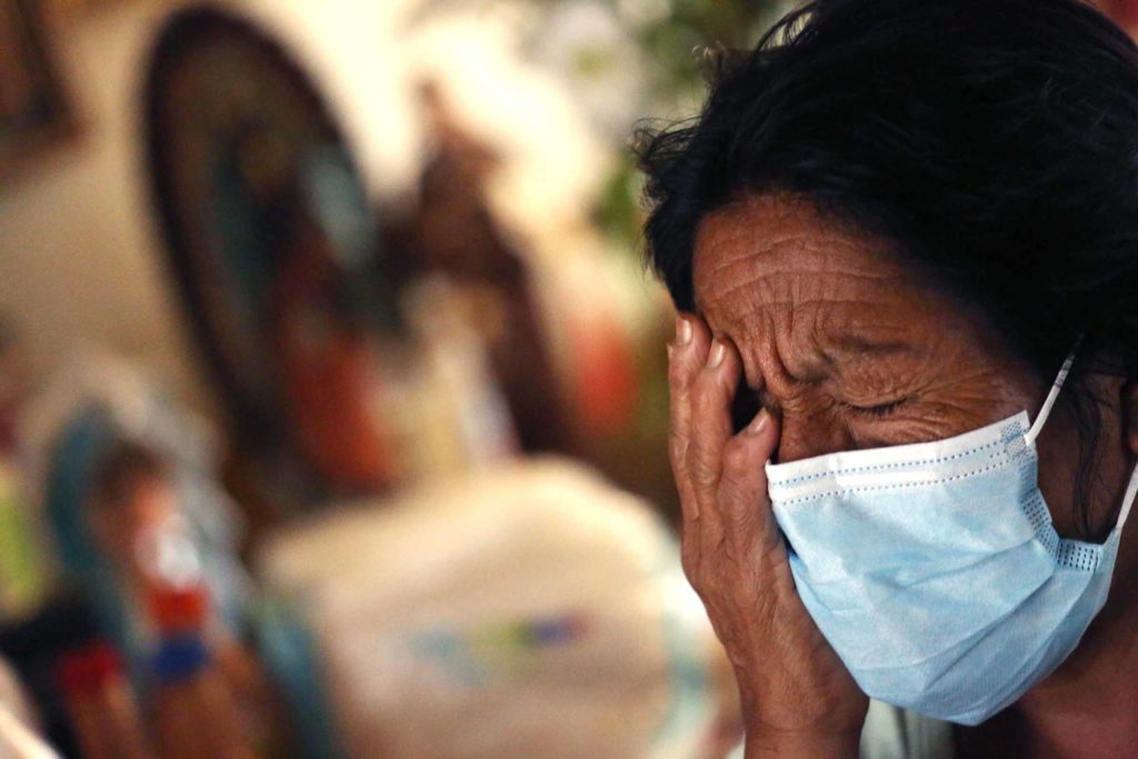 An elderly patient in a medical mask wearily rests her face against her palm