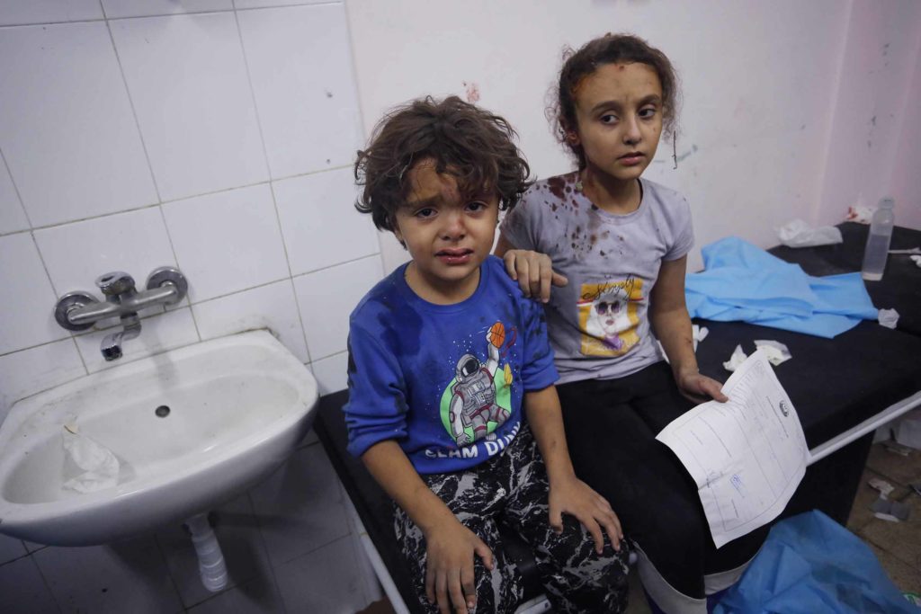 Two injured Palestinian children, one looking directly into the camera, sit on a bench inside of a hospital