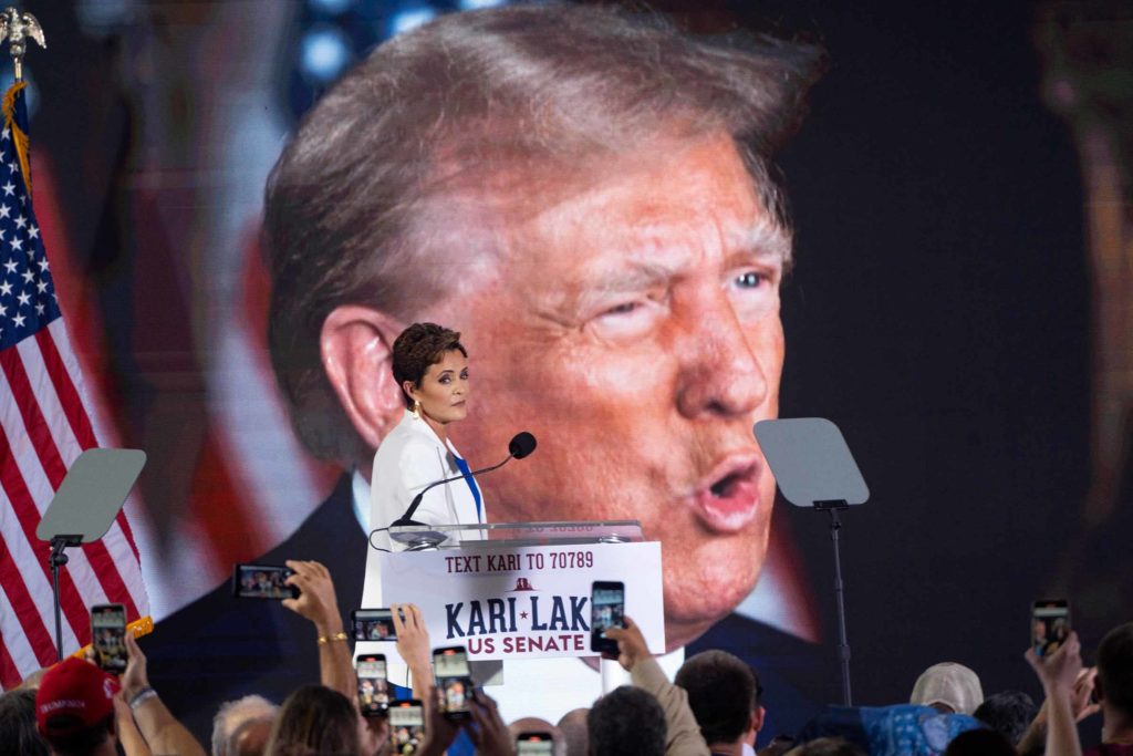 Kari Lake walks in front of a banner of Donald Trump