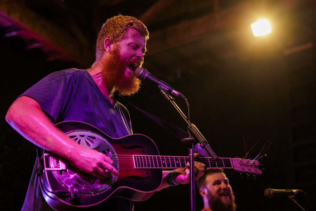 Oliver Anthony is a bearded white man standing in front of microphone playing a guitar bathed in neon light.