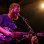 Oliver Anthony is a bearded white man standing in front of microphone playing a guitar bathed in neon light.