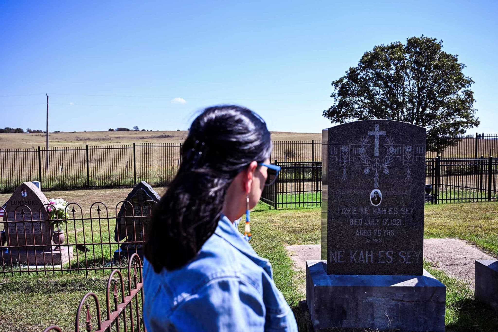 A woman stands in a graveyard, her attention toward the prominently displayed headstone of Lizzie Ne Kah Es Say (1848 - 1921)