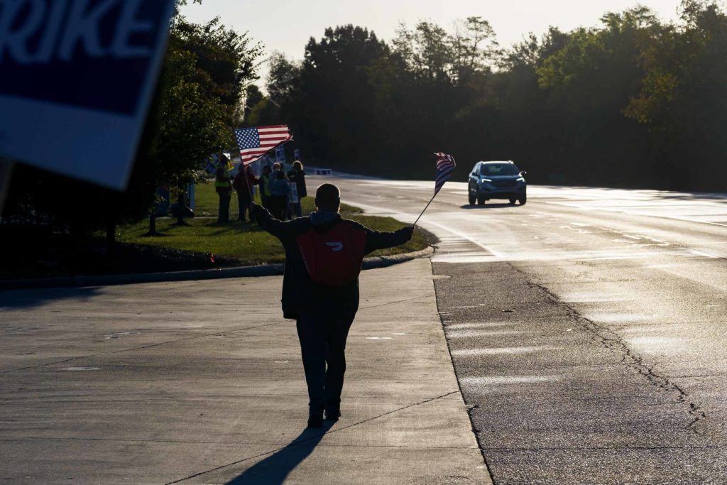 A person holding two small U.S. flags walks towards a group of striking United Auto Workers union members