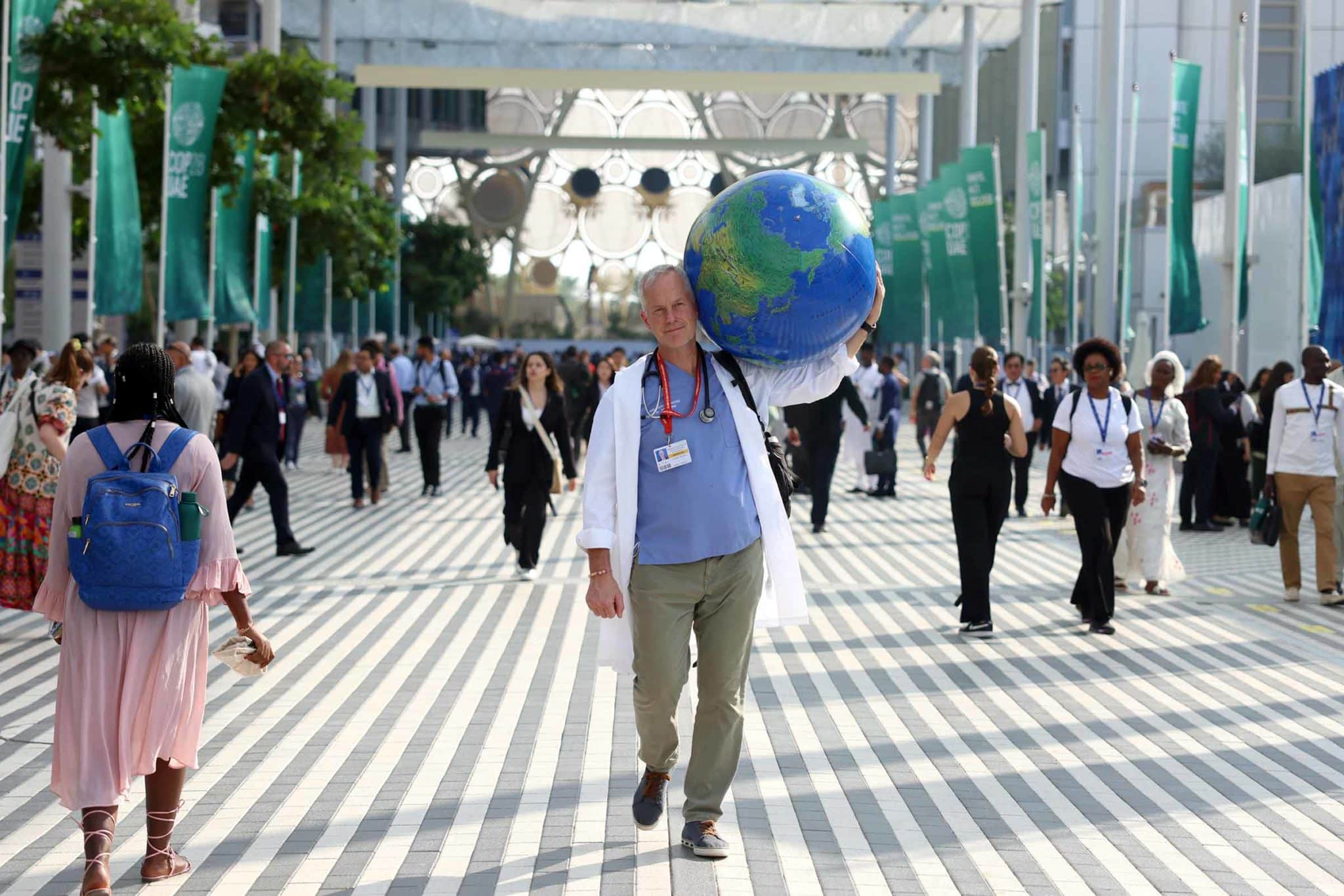 A man wearing a lab coat and stethoscope walks down a hallway carrying an inflated globe