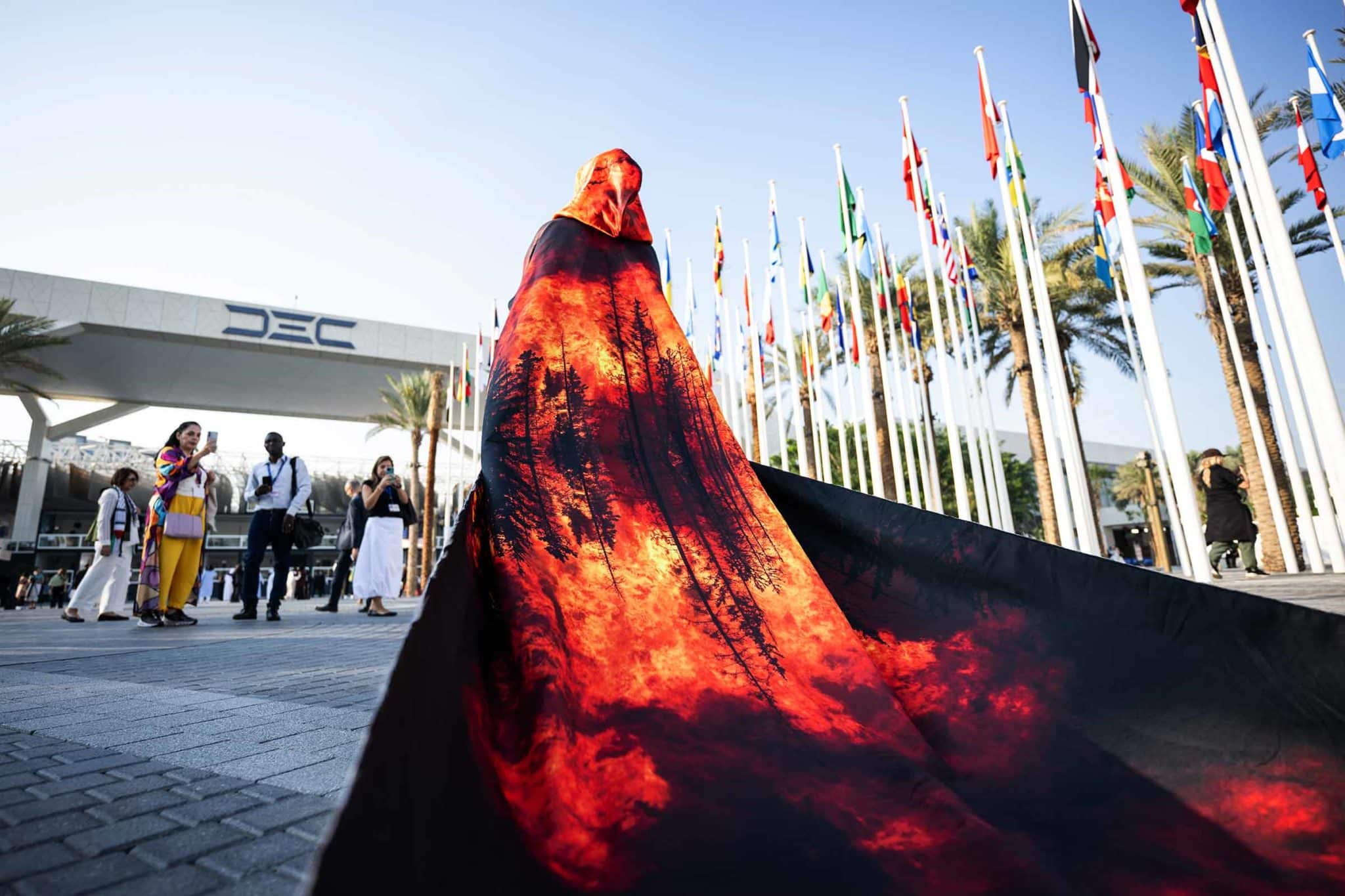 A woman dressed in a cloak printed with the image of a forest burning in a wildfire walks among other participants in COP28