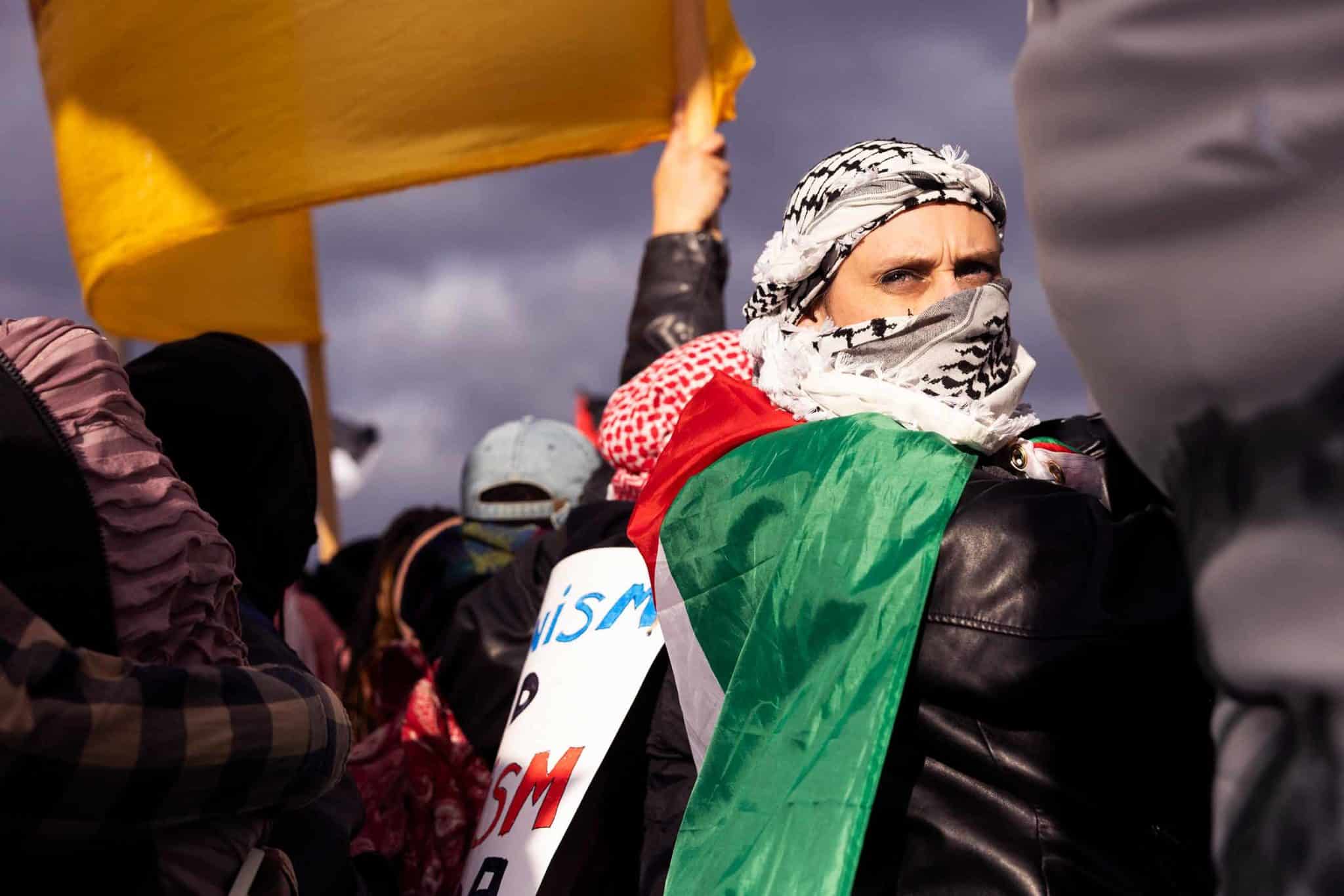 A person draped in the Palestinian flag and whose face is mostly covered with a kuffiyeh stares out into the sun during an outdoor protest