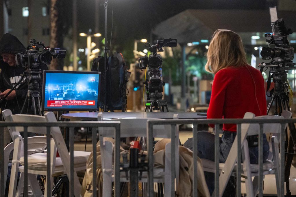 A woman in a red dress sits at a table opposite of several news cameras