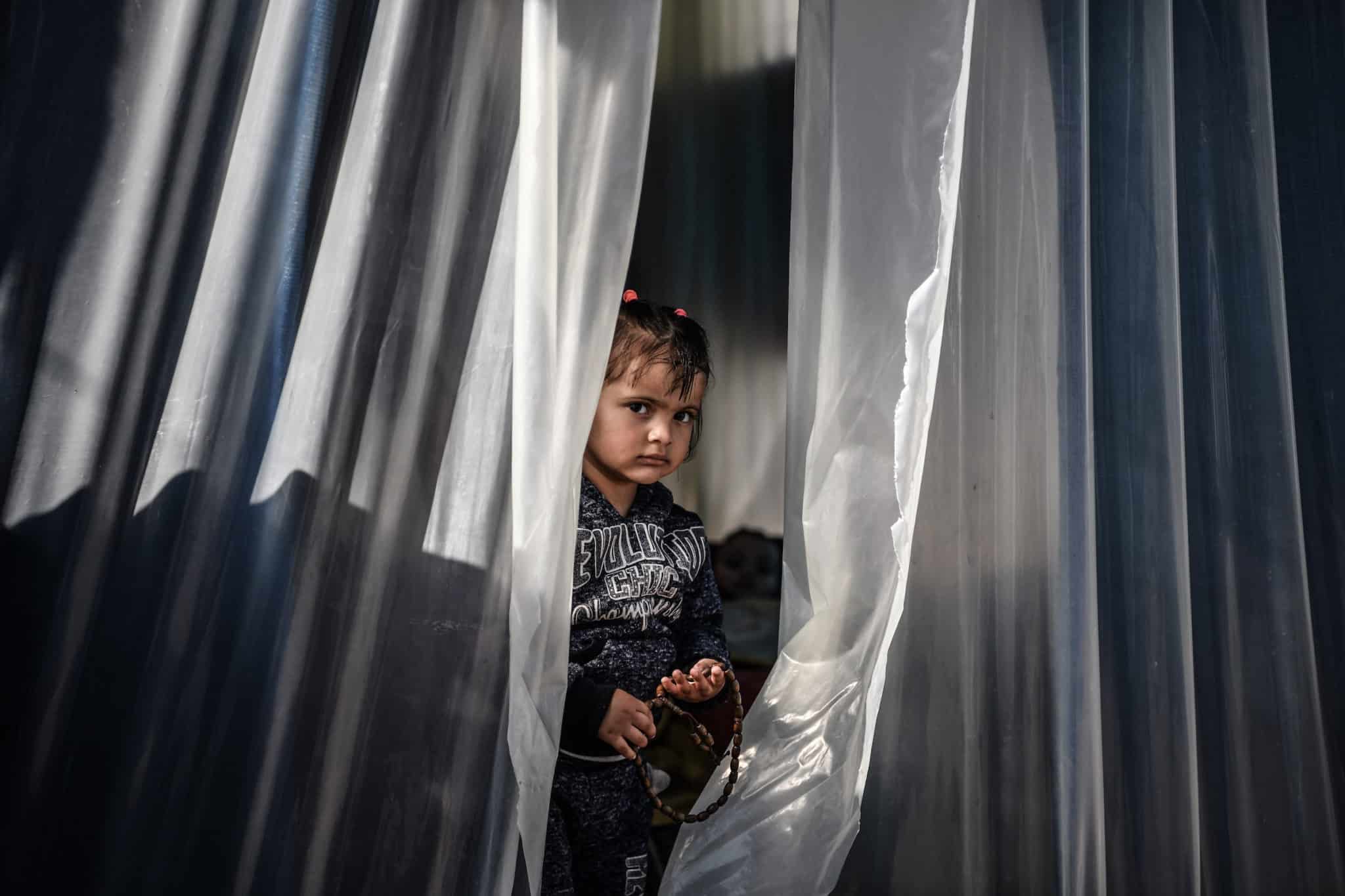 A little girl sits in a makeshift shelter made of a clear plastic curtain.