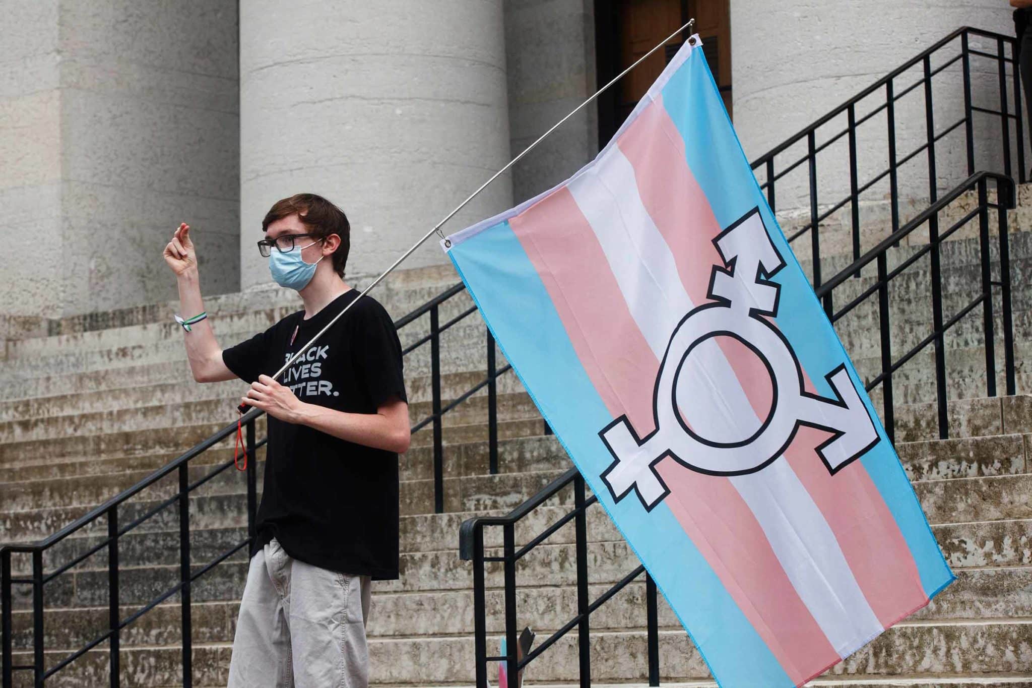 A protester holding the trans flag snaps their fingers while on the steps of a capitol building