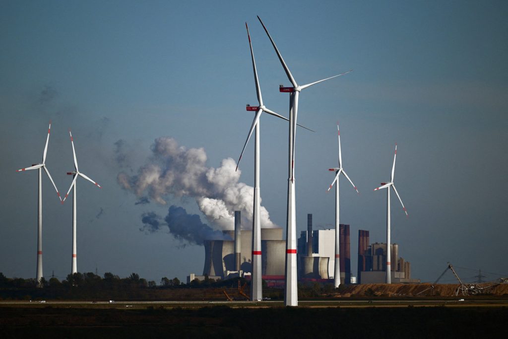 Wind turbines stand in the foreground against smokestacks billowing pollution into the air behind them
