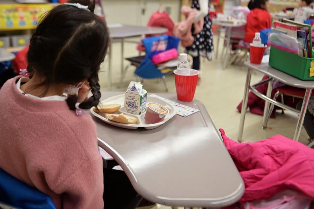 A tiny girl in a tiny chair eats a tiny breakfast with the rest of her tiny kindergarten class