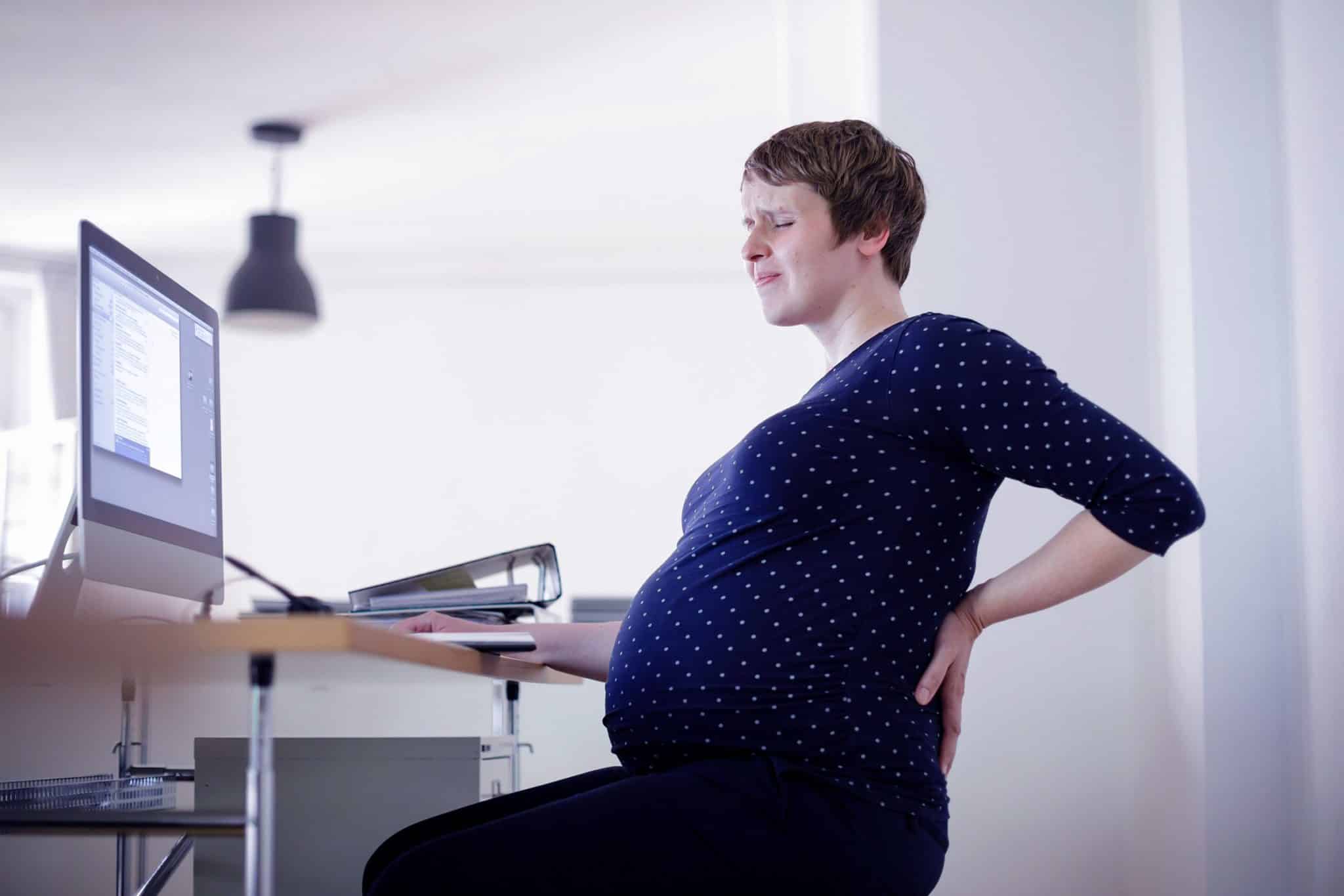 A pregnant person rubs their back while sitting at an office desk